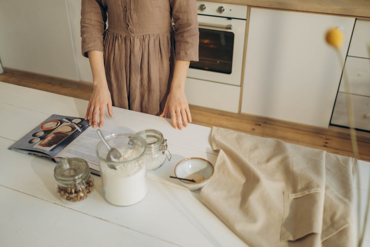 A person prepares ingredients on a wooden kitchen table with a cookbook for baking.